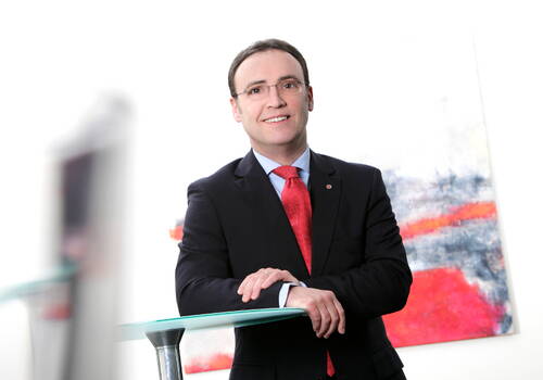 Geschäftsmann im Anzug mit roter Krawatte lehnt lächelnd an einem Glastisch vor einem modernen, abstrakten Gemälde.