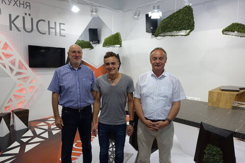 Drei Männer stehen lächelnd in einer modernen Ausstellungsküche mit geometrischem Messestand-Design und hängenden Moos-Elementen im Hintergrund
