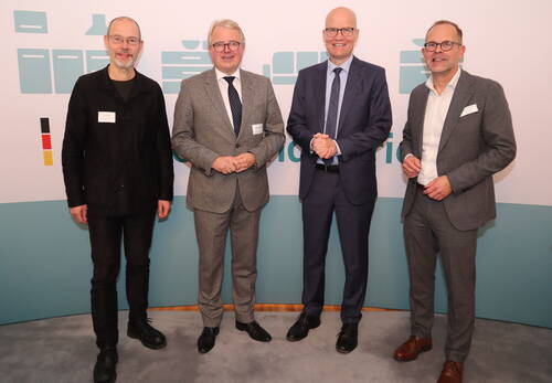 Vier Männer in Anzügen und Namensschildern stehen nebeneinander vor einer hellen Konferenzwand und lächeln in die Kamera