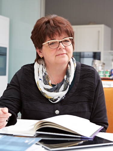 Ältere Frau mit Brille sitzt nachdenklich am Schreibtisch und blättert in einem aufgeschlagenen Buch in moderner Büro- oder Küchenumgebung