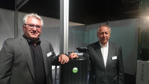 Zwei Männer in Anzugjacken präsentieren an einem Messestand ein schwarzes technisches Gerät mit grünem Logo und lehnen sich lächelnd daran an