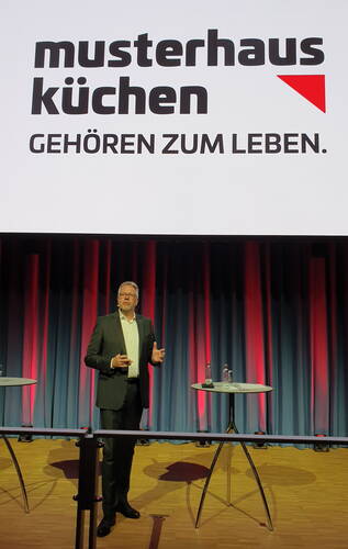 Geschäftsmann hält Präsentation auf einer Bühne vor großem „musterhaus küchen – gehören zum Leben“-Markenlogo mit farbig beleuchtetem Vorhang im Hintergrund