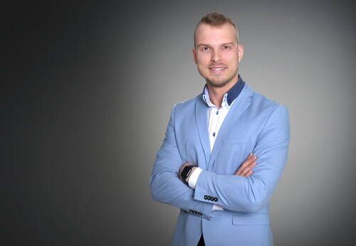 Junger Mann im hellblauen Sakko mit verschränkten Armen lächelt selbstbewusst vor neutral grauem Hintergrund.