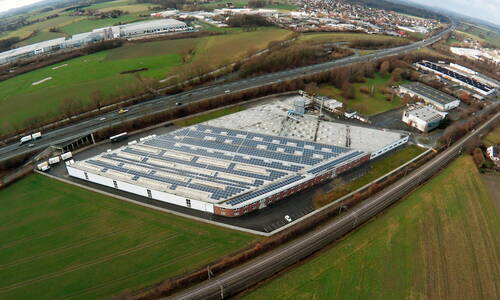 Großes Industriegebäude mit vollständig mit Solarpaneelen bedecktem Flachdach, umgeben von Feldern, Autobahn und Bahnstrecke in einer ländlichen Umgebung