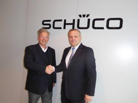 Zwei Geschäftsleute im Anzug geben sich vor einer Wand mit dem Schüco-Logo die Hand und blicken in die Kamera