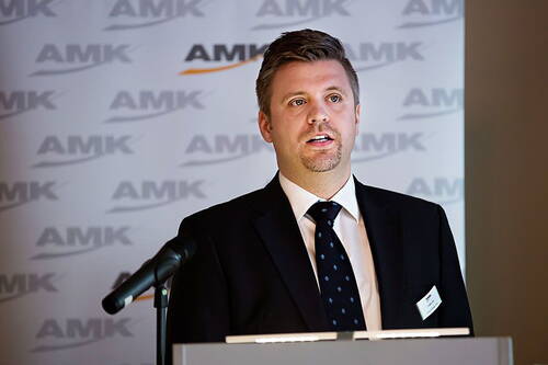 Geschäftsmann im Anzug hält eine Präsentation am Rednerpult vor einer AMK-Branded-Wand und spricht in ein Mikrofon.