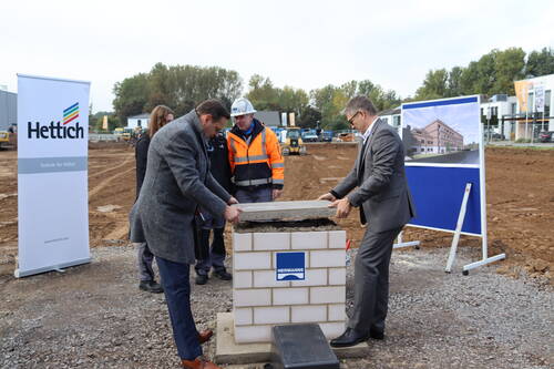 Zwei Männer in Anzug und Mantel setzen gemeinsam eine Betonplatte auf einen Grundstein, während Bauarbeiter und Werbetafeln des Unternehmens Hettich auf der Baustelle im Hintergrund zu sehen sind.