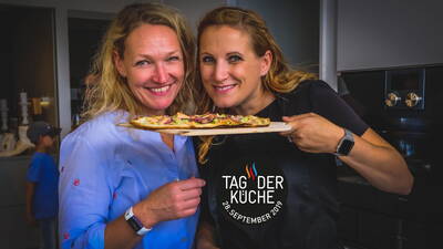 Zwei lachende Frauen präsentieren in einer modernen Küche einen frisch gebackenen Flammkuchen auf Holzbrett beim Event „Tag der Küche“ am 28. September 2019.