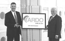 Zwei Geschäftsmänner im Anzug präsentieren in einer Lagerhalle ein großes Schild mit dem ARDO-Logo und dem Slogan „made for you“