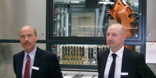 Zwei Männer im Anzug präsentieren eine automatisierte Industrieanlage mit Roboterarm hinter einer Glasscheibe
