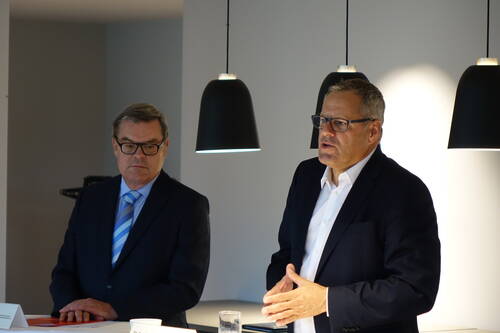 Zwei Geschäftsleute im Anzug stehen unter modernen Hängelampen, einer spricht gestikulierend, der andere hört an einem Konferenztisch aufmerksam zu.