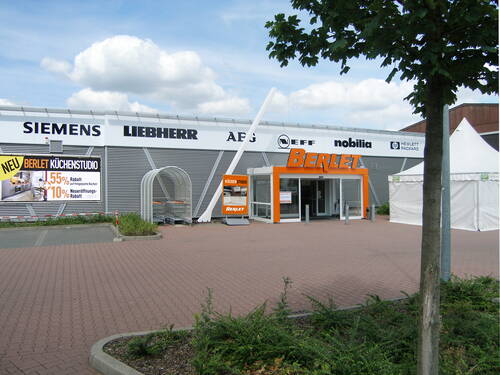 Eingangsbereich eines Berlet-Elektromarkts mit orangem Vordach, Werbeplakat für ein neues Küchenstudio und Einkaufswagenbereich auf einem gepflasterten Parkplatz unter bewölktem Himmel