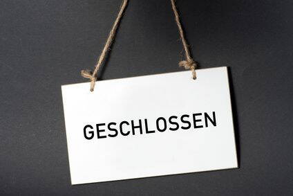 Weißes Schild mit der Aufschrift „Geschlossen“, das an einer Kordel vor dunklem Hintergrund hängt