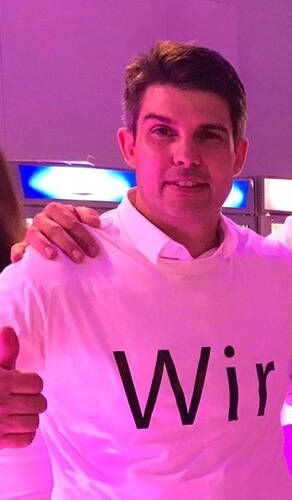 Mann mit weißem T‑Shirt mit dem Aufdruck „Wir“ lächelt in pink beleuchteter Umgebung und zeigt den Daumen nach oben.