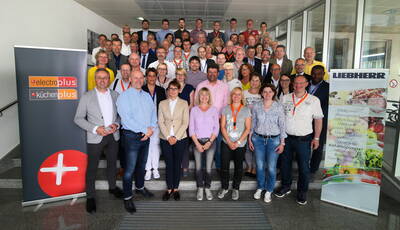 Große Gruppe von Mitarbeiterinnen und Mitarbeitern der Marken electroplus, kuechenplus und Liebherr posiert gemeinsam auf einer Treppe in einem modernen Firmengebäude während einer Veranstaltung.