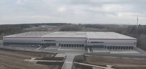 Großes, modernes Logistikzentrum mit zahlreichen Laderampen vor bewaldeter und leicht hügeliger Umgebung unter bedecktem Himmel