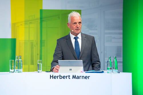 Älterer Mann im Anzug spricht mit Headset-Mikrofon an einem modernen Rednerpult mit Laptop und Wasserflaschen vor farbig beleuchtetem Hintergrund.