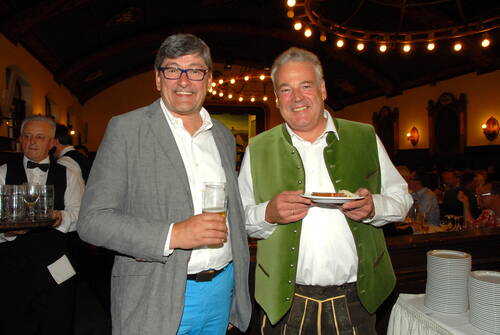 Zwei lachende Männer in festlicher Trachten‑ und Abendkleidung genießen Bier und Essen in einem stimmungsvoll beleuchteten Saal mit vielen Gästen im Hintergrund.