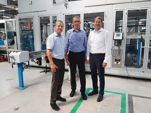 Drei Männer in Businesskleidung stehen lächelnd vor einer großen automatisierten Produktionsanlage in einer modernen Fabrikhalle