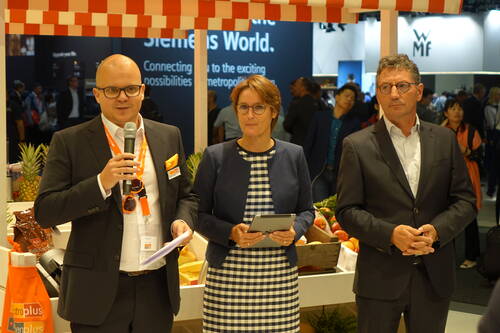 Drei Geschäftsleute in Anzügen präsentieren auf einer Messe an einem Marktstand mit Obst und Gemüse, während der Mann links mit Mikrofon spricht und die Frau in der Mitte ein Tablet hält.