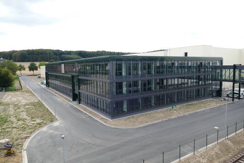 Neues, mehrstöckiges Büro- und Verwaltungsgebäude mit Glasfassade in einem Industriegebiet, umgeben von frisch angelegten Straßen und freier Brachfläche am Waldrand.
