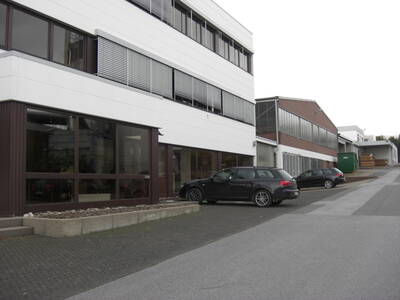 Moderner Büro- und Gewerbekomplex mit großen Fensterfronten, davor geparkte schwarze Kombis auf einer leicht ansteigenden Zufahrtsstraße.