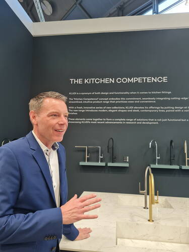 Mann im blauen Anzug erklärt an einem Ausstellungsstand mit der Aufschrift „The Kitchen Competence“ verschiedene moderne Küchenarmaturen an einer hellen Marmorarbeitsplatte