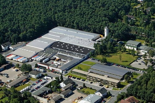 Luftaufnahme eines großen Industrie- und Bürokomplexes am Waldrand mit Lagerhallen, Parkplätzen und angrenzenden Wohnhäusern