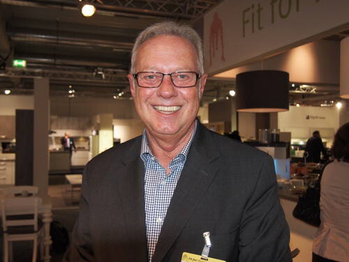 Älterer Mann mit Brille und grauem Haar lächelt in einem modernen Messestand mit Küchen- und Einrichtungselementen im Hintergrund