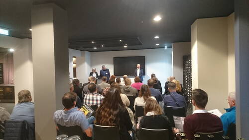 Publikum verfolgt eine Präsentation, bei der drei Vortragende an Pulten vor einem großen Bildschirm in einem modernen Konferenzraum sprechen.