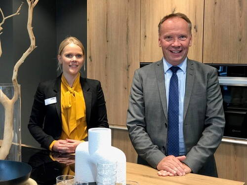 Zwei lächelnde Geschäftsleute stehen hinter einer modernen Holzküchentheke mit Dekovasen und Geschirr vor einem Einbaubackofen.