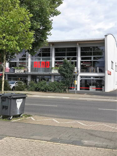 Modernes Geschäftsgebäude mit großen Glasfronten und ALNO-Schriftzug an einer mehrspurigen Straße, im Vordergrund ein Restmüllcontainer und ein Fußweg mit Kopfsteinpflaster.