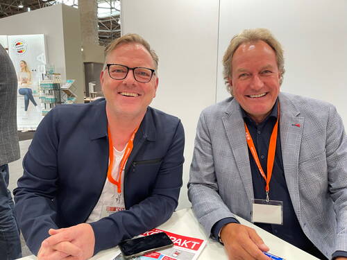 Zwei lächelnde Männer mit orangefarbenen Messeausweisen sitzen nebeneinander an einem Tisch in einer hellen Ausstellungshalle