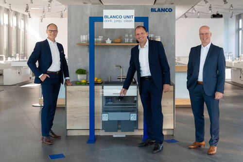 Drei Geschäftsleute in Anzügen präsentieren in einer modernen Ausstellungsküche eine BLANCO UNIT Spülen- und Abfalltrenneinheit unter einem blau gerahmten Display.