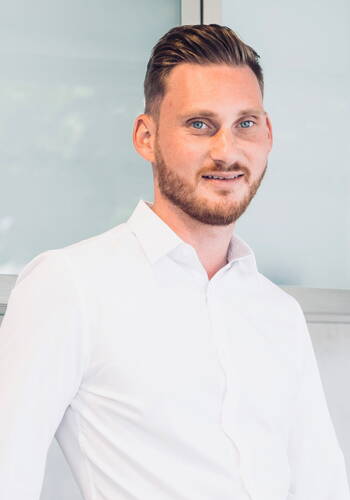 Junger Mann mit Bart und zurückgekämmten Haaren lächelt in weißem Hemd vor hellem, modernem Bürohintergrund