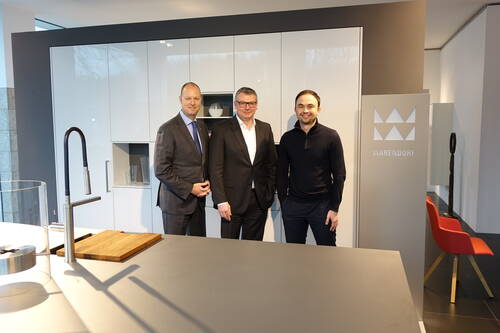Drei Männer posieren in einer modernen Ausstellungs­küche neben einer Kücheninsel mit Spüle vor weißen Hochglanzschränken und einem Warendorf-Logo.
