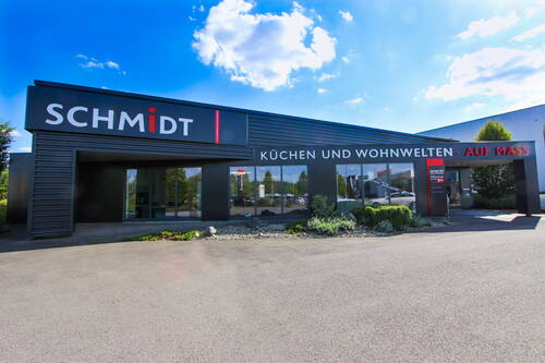 Modernes SCHMIDT-Küchenstudio mit großer Glasfassade und Beschriftung „Küchen und Wohnwelten – auf Maß“ an einem sonnigen Tag