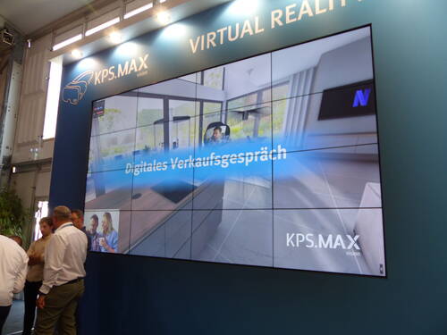 Große Messedisplaywand von KPS.MAX mit der Aufschrift „Digitales Verkaufsgespräch“ zeigt eine virtuelle Wohnraumansicht, während mehrere Besucher davor stehen und sich unterhalten