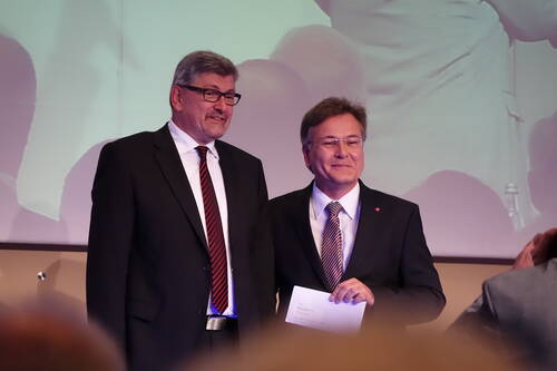 Zwei festlich gekleidete Männer im Anzug stehen lächelnd nebeneinander auf einer Bühne, während einer ein Dokument in der Hand hält und im Hintergrund eine vergrößerte Projektion der Szene zu sehen ist.