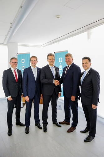 Fünf Geschäftsleute in Anzügen stehen in einem hellen Konferenzraum, zwei von ihnen geben sich in der Mitte der Gruppe die Hand vor blauen Firmenbannern.
