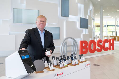 Geschäftsmann in Bosch-Ausstellungshalle präsentiert eine Reihe elektronischer Bauteile und ein weißes Analysegerät auf einem Tresen mit großem roten Bosch-Schriftzug im Hintergrund