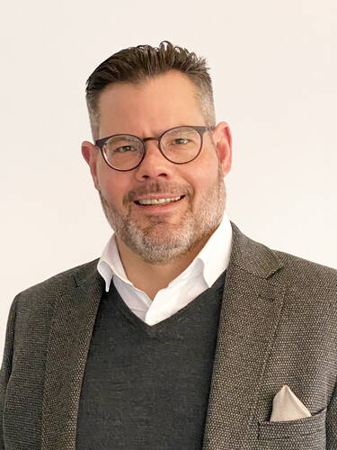 Lächelnder Mann mit Brille und Bart in grauem Sakko und dunklem Pullover vor hellem Hintergrund