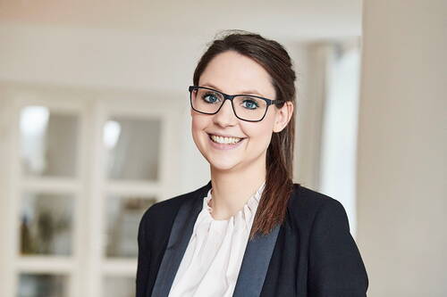 Junge Frau mit Brille und dunklem Blazer lächelt freundlich in einem hellen Büro mit unscharfem Hintergrund