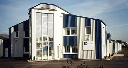 Modernes zweistöckiges Industriegebäude mit blau-weißer Metallfassade, großem verglastem Treppenhaus und klar sichtbarem Firmenschild an der Frontseite