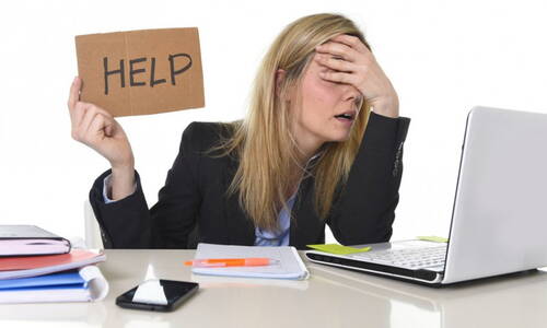 Gestresste Büroangestellte sitzt erschöpft vor dem Laptop, hält sich die Hand vors Gesicht und ein Pappschild mit der Aufschrift „HELP“ hoch