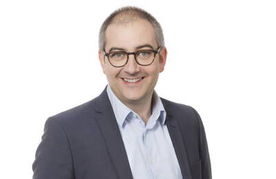 Lächelnder Mann mit Brille in Hemd und dunklem Anzug vor weißem Hintergrund