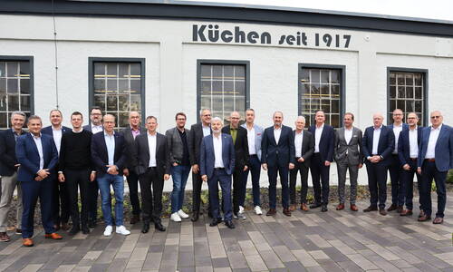 Gruppe von Geschäftsleuten in Anzügen steht vor einem historischen Firmengebäude mit der Aufschrift „Küchen seit 1917“.
