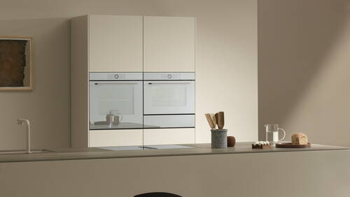 Minimalistische beige Küche mit Doppelbackofen, Kochinsel, Holzutensilien, Wasserkrug und angeschnittenem Brotlaib auf der Arbeitsplatte