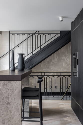 Minimalistische Küche mit grauer Steininsel, schwarzem Barhocker und Vasen vor einer modernen Metalltreppe mit grafischer Wandgestaltung