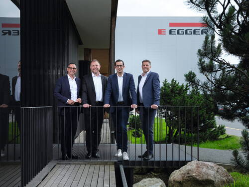 Vier Geschäftsleute in Anzügen stehen lächelnd auf einer kleinen Metallbrücke vor einem modernen Firmengebäude mit EGGER-Logo und umgebender Grünanlage.